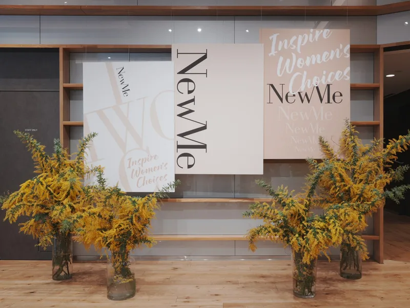 「Inspire Women's Choices」と書かれた NewMe のポスターの前に、ミモザの花が飾られている様子